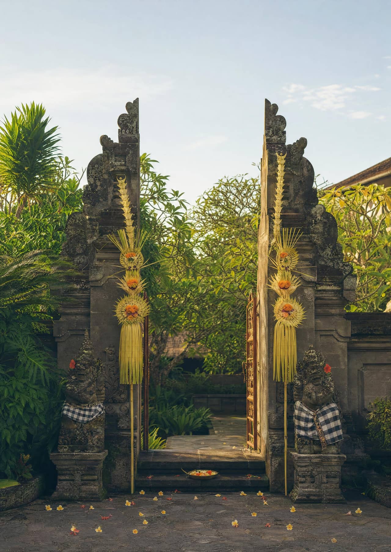 A Balinese welcome at a hotel entrance in warm morning light: a carved stone gateway hung with woven palm leaf, a stone guardian statue in checked poleng cloth, and frangipani flowers scattered on dark paving.